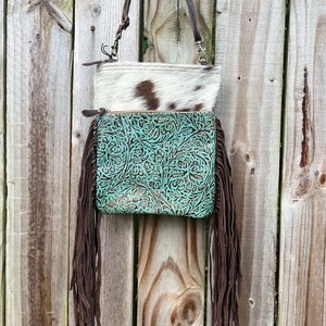 May include: A two-tone crossbody bag with a cowhide top and a turquoise leather bottom. The turquoise leather is embossed with a floral pattern and has long brown fringe. The bag has a brown leather strap.