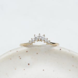 May include: A delicate gold ring featuring a curved design adorned with multiple clear, sparkling gemstones. The ring's band is thin, and the gemstones are arranged in a gentle arc, creating a subtle yet elegant aesthetic. The ring is displayed on a speckled white surface.