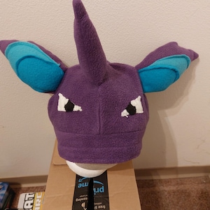 May include: A purple fleece hat with a central horn, blue ear accents, and white eye details. The hat is designed to resemble a fictional creature. The hat is displayed on a white head form, placed on a cardboard box.