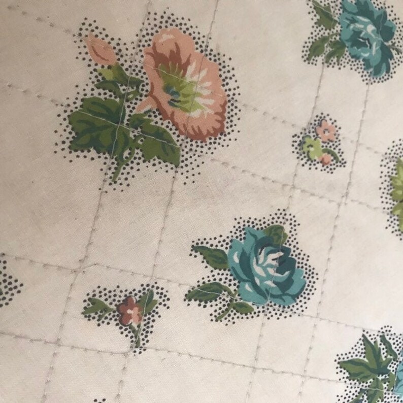 May include: A white fabric with a quilted pattern and a floral print. The flowers are in shades of pink, blue, and green.