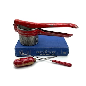 May include: Red metal potato masher with a handle and a strainer. The masher is sitting on top of a blue book titled "The Presidents' Cookbook".