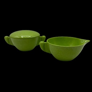 May include: A two-piece green serving set. The set includes a lidded bowl and a creamer with a spout. Both pieces have a textured surface and small handles. The set is made of a light green material.