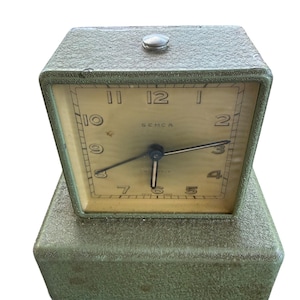 May include: A vintage Semca table clock with a textured olive green metal casing. The square clock face features a cream background with black Arabic numerals, thin black hands, and a small silver button on the top of the housing. The base is rectangular and matches the textured green finish of the clock body.