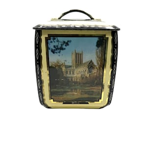 May include: A rectangular metal tin with a hinged lid and handle, featuring a black and cream border with a decorative white diamond pattern. The front panel displays a color illustration of Wells Cathedral with the text WELLS printed below.