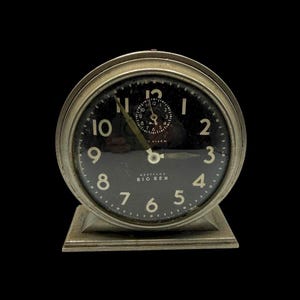 May include: A vintage Westclox Big Ben alarm clock with a round silver-toned metal casing and a square base. The clock face is black with white Arabic numerals, a smaller alarm dial at the top, and thin metallic hands indicating the time.