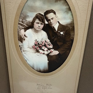 May include: Vintage sepia-toned photograph of a couple in formal attire. The woman wears a white dress and pearl necklace, holding a bouquet. The man is in a dark suit. The photo is in an oval frame with the studio name and address printed below.