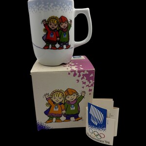 May include: White ceramic mug with a handle, featuring a cartoon illustration of two children in winter clothes. The mug is on a box with a matching design. Includes a tag with the Lillehammer '94 Olympic logo.