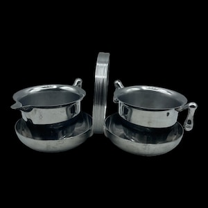 May include: A silver serving set with two small bowls and a handle. The bowls have a rounded shape and a small spout. The handle is a long, thin piece of metal that connects the two bowls.