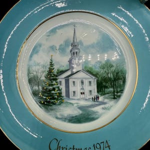 May include: A decorative plate with a teal rim and a gold trim. The center features a painted scene of a white church with a tall steeple, a Christmas tree, and the text "Christmas 1974".