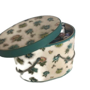 May include: A vintage oval-shaped storage box with a teal lid and a floral pattern. The box has a rope handle and is filled with spools of thread.