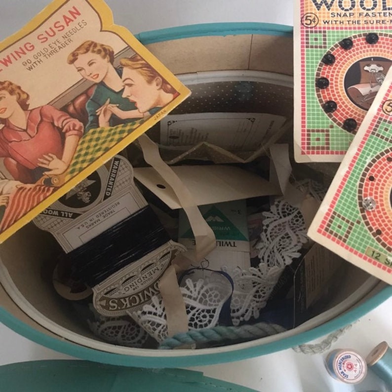 May include: A turquoise blue round tin box filled with various sewing supplies, including lace, thread, buttons, and a card that reads "Sewing Susan Do Gold Eye Needles with Threader".