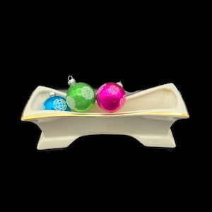 May include: A white ceramic Christmas tree ornament holder with a gold trim. Three glass ornaments are inside the holder: one blue, one green, and one pink.