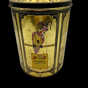May include: A gold metal tin with a black lid. The tin has a colorful illustration of a bird with a feathered hat and a yellow necklace. The text on the tin reads "Electrifying Ensemble Gilded Cage Five Singing Birds High Voltage Always".