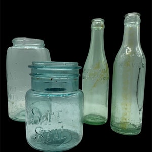 May include: Four vintage glass bottles, two with a light blue tint and two with a clear glass tint. The bottles are all different shapes and sizes. One bottle has the text "Safe Seal" on the side.