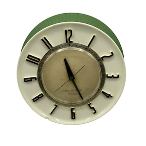 May include: Vintage General Electric wall clock with a white face, black numerals, and a green backing. The clock has a circular design with a glass cover and a silver-colored bezel. The hands point to 5:58.