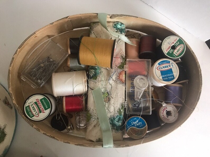 May include: A wooden sewing box filled with various sewing supplies, including spools of thread in different colors, needles, pins, and a thimble. The box also contains a small piece of fabric with a floral pattern.