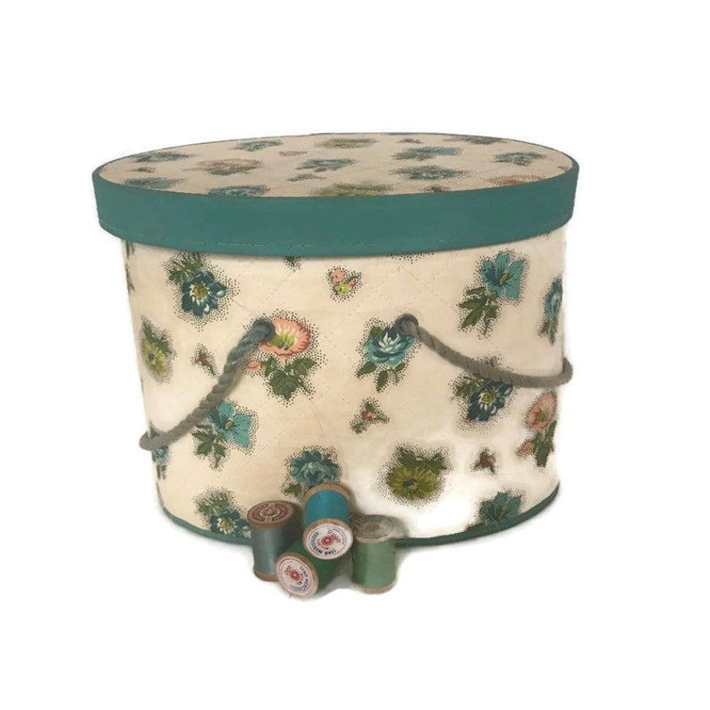 May include: A vintage round hat box with a floral pattern and teal trim. The box has rope handles and is sitting on a white surface with spools of thread.