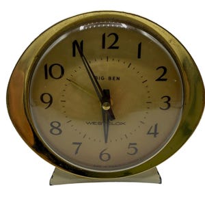 May include: A round Westclox Big Ben alarm clock with a gold-toned metal frame and a matching gold-colored face. The clock features black Arabic numerals from one to twelve, black hour and minute hands, a thin second hand, and a clear glass cover.