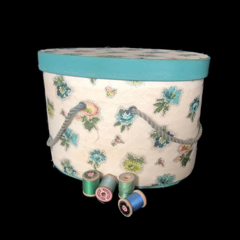 May include: A vintage oval-shaped hat box with a turquoise lid and a floral pattern. The box has a rope handle and is sitting on a surface with spools of thread in various colors.