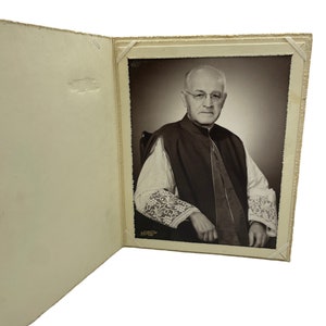 May include: A black and white photograph of a man wearing a clerical collar and a cassock. He is sitting in a chair and has his hands clasped in his lap. The photo is framed in a white mat and is mounted in a white album.