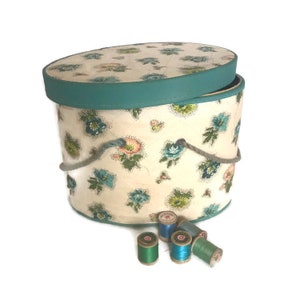 May include: A vintage oval-shaped hat box with a floral print and a teal lid. The box has a rope handle and is open, revealing the interior. There are several spools of thread in various shades of blue and green sitting in front of the box.