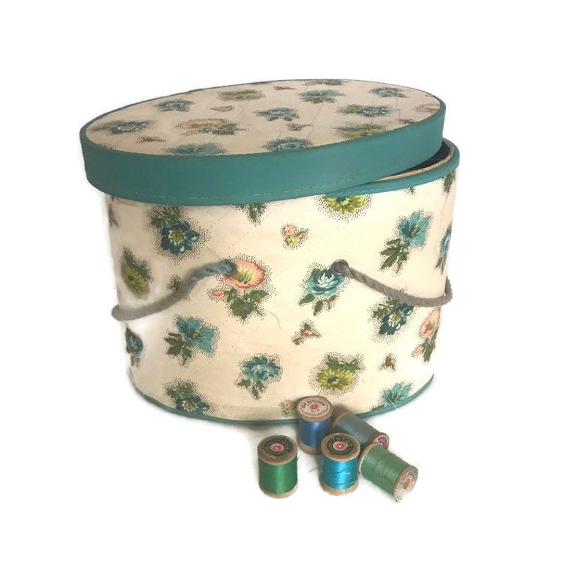 May include: A vintage oval-shaped hat box with a floral print and a teal lid. The box has a rope handle and is open, revealing the interior. There are several spools of thread in various shades of blue and green sitting in front of the box.