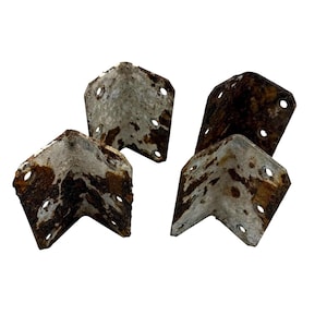 May include: Four vintage metal corner braces with a distressed, rusty appearance. Each L-shaped bracket has multiple pre-drilled holes for mounting. The metal shows signs of age and wear, with a mix of silver and brown tones.