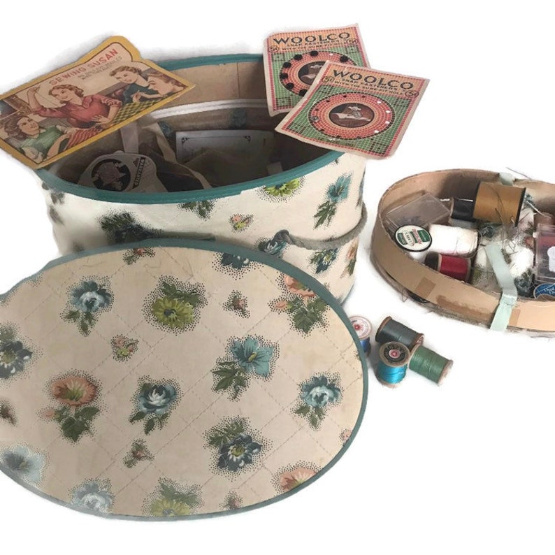 May include: A vintage floral hat box with a blue trim and a lid. The box is filled with various sewing supplies, including spools of thread, buttons, and needles. The box is sitting on a white surface.