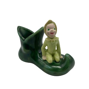 Könnte beinhalten: Vintage Keramikfigur eines sitzenden Elfen in grünem Outfit, der auf einem grünen stiefelförmigen Pflanzgefäß sitzt. Der Elf hat ein lächelndes Gesicht und trägt einen spitzen Hut. Der Pflanztopf ist glänzend dunkelgrün.