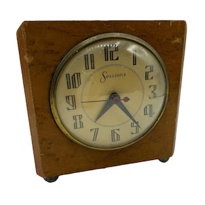 May include: A vintage Sessions clock with a square wooden frame. The clock face is cream-colored with black numerals and hands. The word "Sessions" is printed on the face. The clock has a brass-colored bezel and small feet.