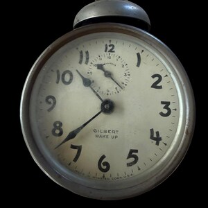 May include: A vintage round metal alarm clock with a cream-colored face, black numerals, and a small sub-dial. The clock features the text GILBERT WAKE UP and WINSTED, CONN. U.S.A. A white cotton swab rests on top of the metal bell at the top of the clock.