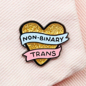 May include: A heart-shaped enamel pin with a gold glitter center and black outline. The pin features two banners with the words "NON-BINARY" and "TRANS" in black text. The banners are light blue and pink. The pin is set against a pink fabric.
