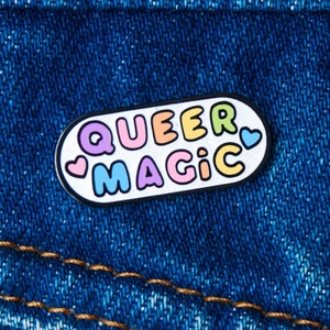 Könnte beinhalten: Eine weiße Emaille-Anstecknadel mit einem Regenbogen-Umrandung und dem Text "Queer Magic" in einer Regenbogen-Schriftart. Die Anstecknadel befindet sich auf einer blauen Jeansjacke.