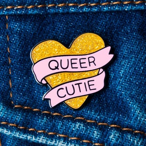 Peut inclure: Une épingle en forme de cœur en paillettes dorées avec une bannière rose qui dit "Queer Cutie".