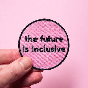 May include: A circular embroidered patch with a bright pink background and a black stitched border. The text "the future is inclusive" is embroidered in black, lowercase, sans-serif lettering across the center of the circular fabric patch.