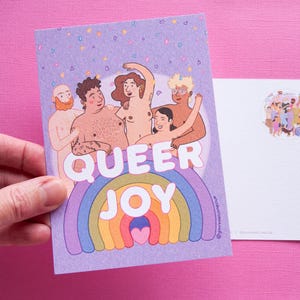 Peut inclure: Une carte postale avec les mots "QUEER JOY" au-dessus d'un motif arc-en-ciel. La carte postale présente une illustration de plusieurs personnes s'embrassant. Un autocollant avec un groupe de personnes est à droite.
