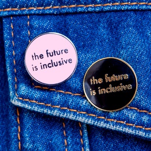 Może przedstawiać: Dwa okrągłe piny z tekstem "the future is inclusive" na niebieskim dżinsowym tle. Pin po lewej stronie jest różowy z czarnym tekstem. Pin po prawej stronie jest czarny z złotym tekstem.