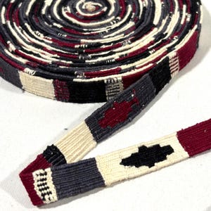 May include: A coiled length of woven fabric trim featuring a repeating geometric pattern in deep burgundy, charcoal grey, black, and off-white. The flat, narrow textile strip displays a consistent, tight weave with alternating colour blocks.