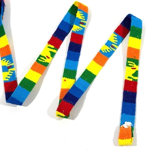 May include: A woven fabric ribbon featuring a repeating pattern of horizontal rainbow stripes in red, orange, yellow, green, and blue, interspersed with yellow sun-shaped motifs. The ribbon is coiled at one end and extends outward in a zigzag.