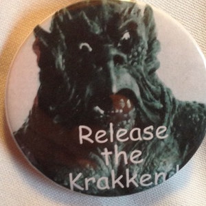 Puede incluir: Un botón redondo con un monstruo verde y negro con una sonrisa de dientes. El texto "Release the Krakken" está escrito en blanco en el botón.