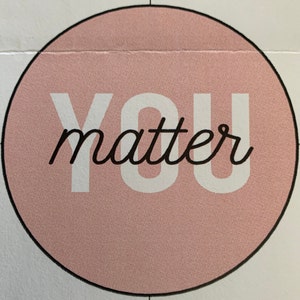 May include: A circular pink graphic featuring the words "YOU matter" in white and black. The text is centrally positioned within the pink circle, which is outlined in black. The background is white with a grid pattern.