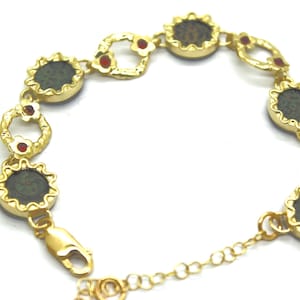 Puede incluir: Pulsera de oro con piedras redondas de color verde oscuro engastadas en marcos de oro. La pulsera tiene pequeñas piedras rojas entre cada piedra. La pulsera tiene un cierre.