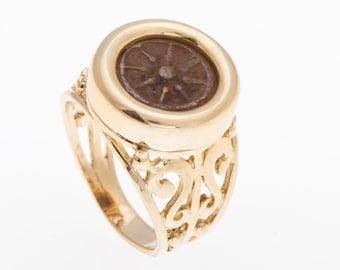 May include: A gold ring with a round, ornate setting featuring a brown coin with a sun-like design. The ring band is made of delicate, intricate gold filigree.