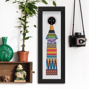 May include: A framed cross-stitch artwork featuring a stylized figure with a black head and a body composed of stacked, patterned sections in various colors. The artwork is in a black frame, and the overall design is geometric and colorful.