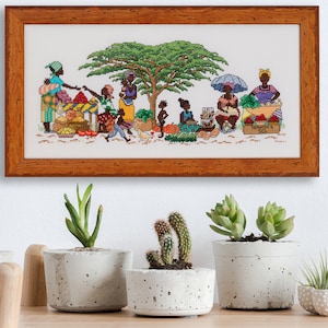Könnte beinhalten: Ein gerahmtes Kreuzstich-Kunstwerk, das eine farbenfrohe afrikanische Marktszene mit Menschen zeigt, die Produkte und Waren verkaufen. Das Kunstwerk wird an einer weißen Wand über einem Regal mit Topfpflanzen ausgestellt.