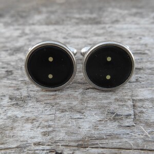 May include: A pair of silver cufflinks with black circular insets and two small yellow dots on each inset.