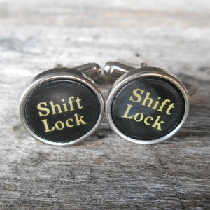 May include: A pair of silver cufflinks with black round buttons. The buttons have the words "Shift Lock" printed in gold letters.
