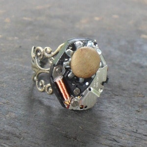 May include: A steampunk-style ring featuring a watch movement with a gold-toned button and a decorative filigree band.