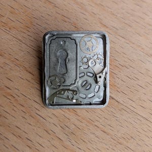 May include: A silver metal keyhole charm with a keyhole design and various gears and cogs around it.