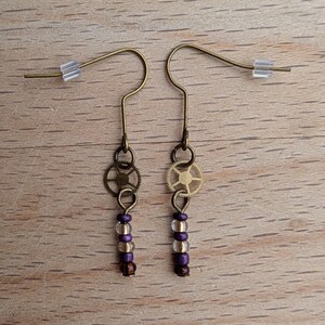 May include: A pair of dangle earrings featuring bronze-toned metal ear wires with clear plastic backings. Each earring includes a small circular gear charm suspended above a vertical arrangement of three beads in alternating shades of purple and clear.